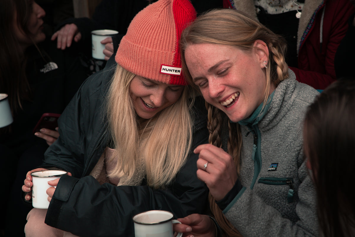 Two women laughing together with enamel mugs at a Grounded Connections meet-up
