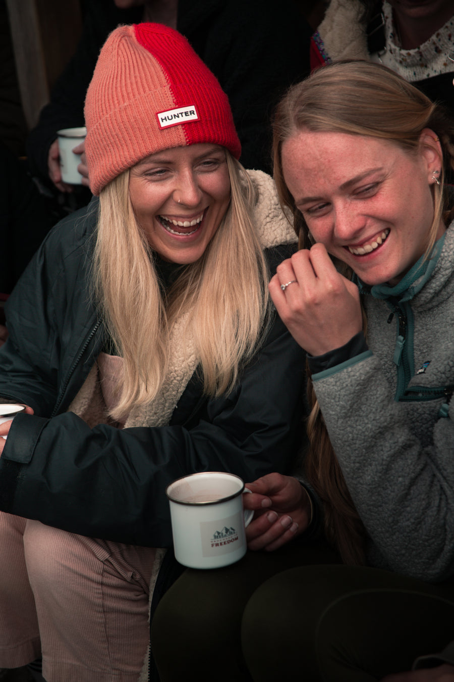 Two women laughing together with enamel mugs at a Grounded Connections meet-up