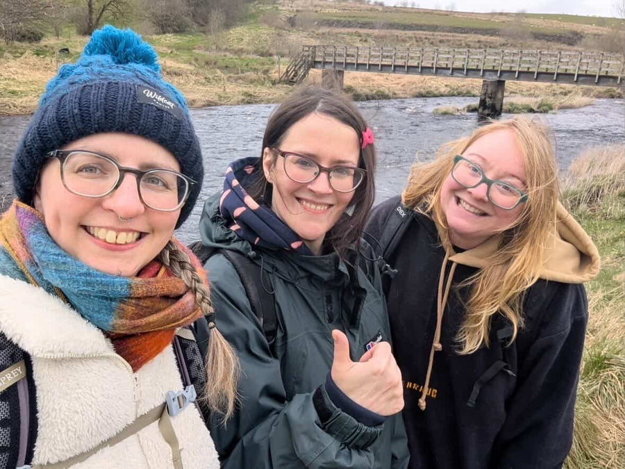 Three women friends smiling by a river on a Grounded Connections walk